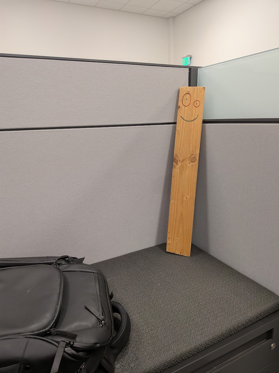 A wooden plank with a smiley face drawn on it, leaning in a cubicle corner
