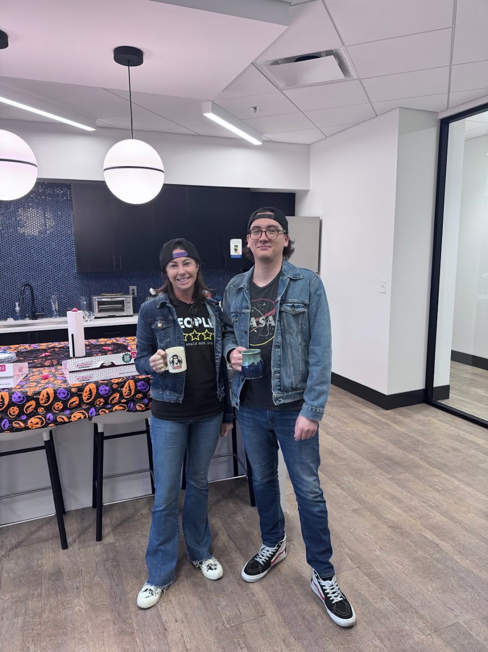 Two people in an office kitchen, both wearing backwards caps and denim jackets; Alex on the right, a product teammate on the left in a matching outfit